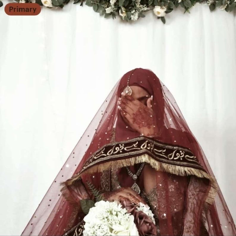 Person wearing a red veil with intricate designs, holding flowers against a white background.