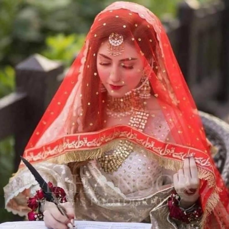 Woman in traditional attire with a red veil, sitting and writing on a piece of paper.